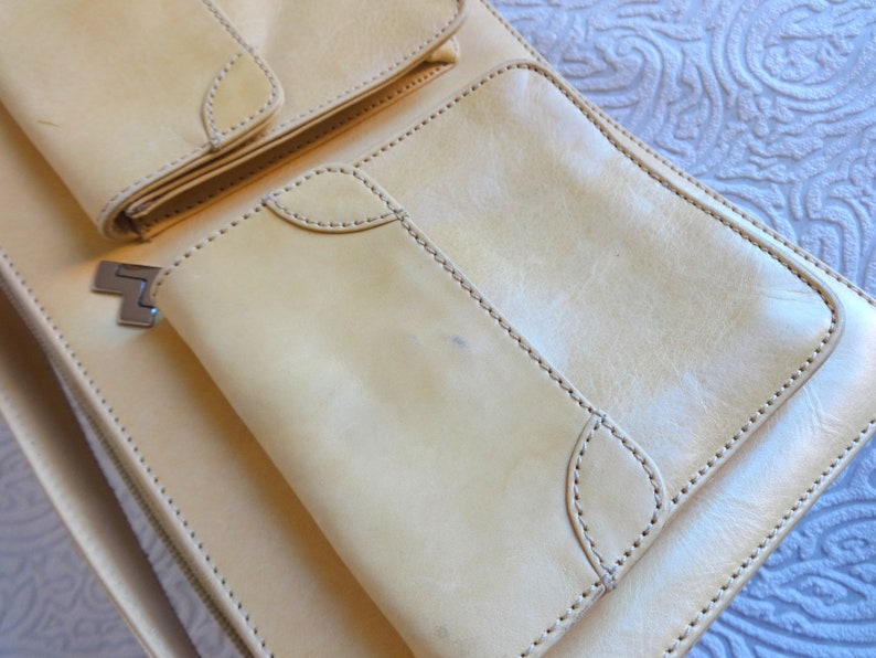 May include: A light beige leather bag with multiple pockets and a zipper closure. The bag has a flap with a metal clasp.