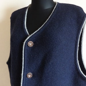 Puede incluir: Un chaleco de punto azul marino con ribete gris alrededor del cuello y las sisas. El chaleco tiene cuello en V y tres botones en la parte delantera.