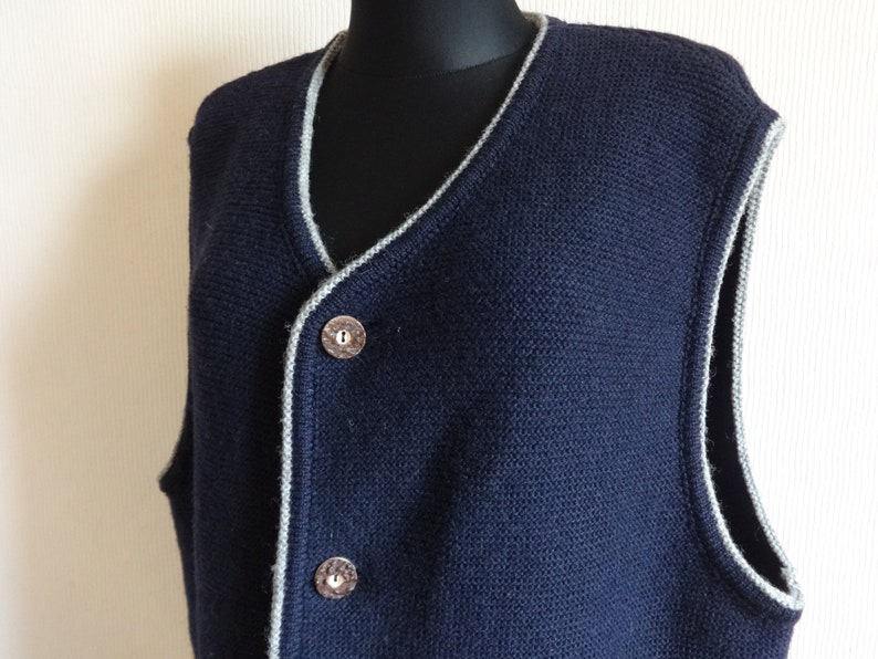 Puede incluir: Un chaleco de punto azul marino con ribete gris alrededor del cuello y las sisas. El chaleco tiene cuello en V y tres botones en la parte delantera.