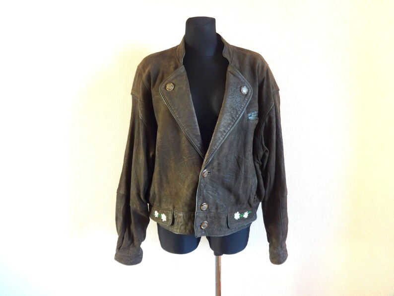 Puede incluir: Una chaqueta de cuero marr&oacute;n con cierre de cremallera y detalles de botones. La chaqueta tiene cuello y mangas largas. Hay flores blancas bordadas en la parte inferior de la chaqueta.