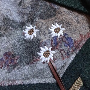 Puede incluir: Un su&eacute;ter verde con una correa de cuero marr&oacute;n y tres flores de edelweiss blancas bordadas sobre un fondo gris.