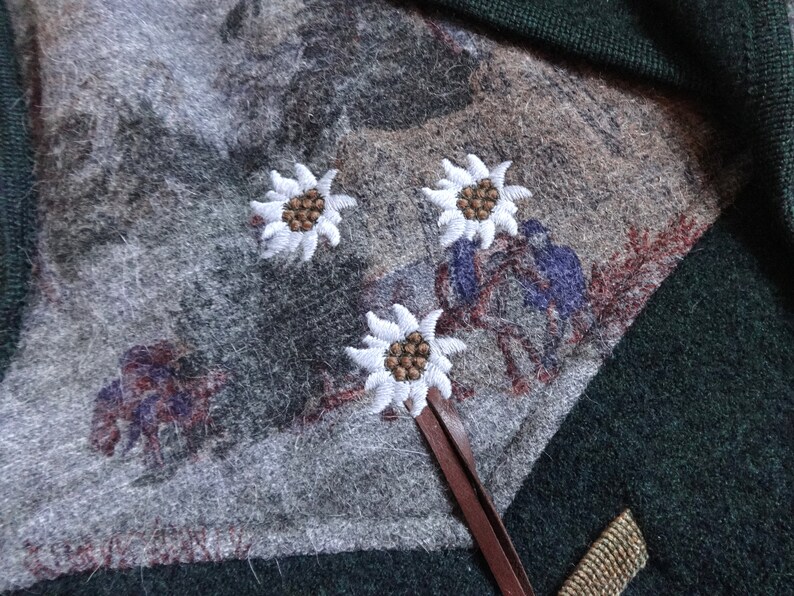 Puede incluir: Un su&eacute;ter verde con una correa de cuero marr&oacute;n y tres flores de edelweiss blancas bordadas sobre un fondo gris.