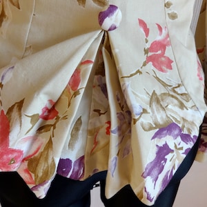 Puede incluir: Primer plano de una chaqueta floral beige con un dise&ntilde;o plisado. La chaqueta presenta un bot&oacute;n grande y redondo y est&aacute; adornada con estampados florales rosas, morados y marrones. Las mangas son visibles a los lados.