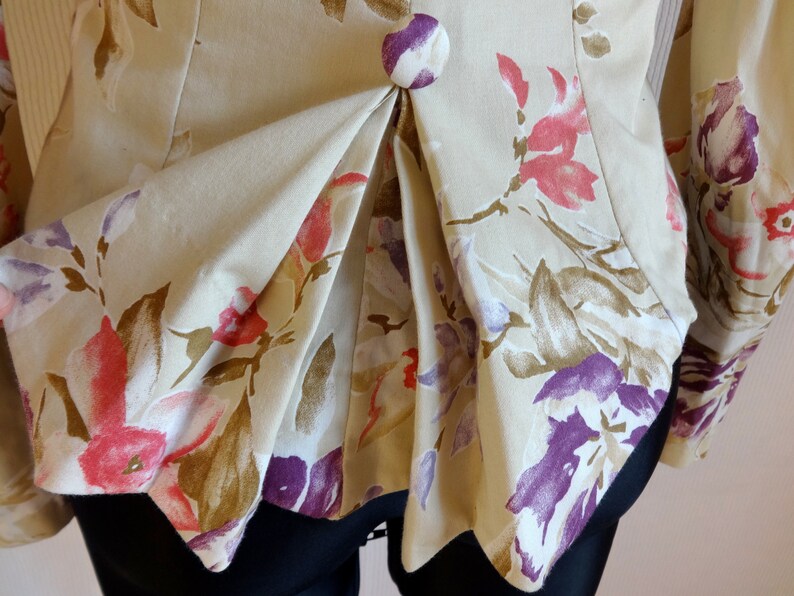 Puede incluir: Primer plano de una chaqueta floral beige con un dise&ntilde;o plisado. La chaqueta presenta un bot&oacute;n grande y redondo y est&aacute; adornada con estampados florales rosas, morados y marrones. Las mangas son visibles a los lados.