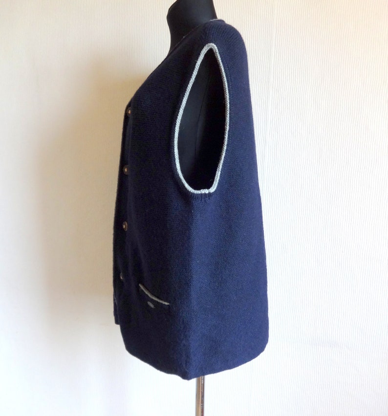 Puede incluir: Un chaleco de punto azul marino con ribete gris claro alrededor de las sisas y el escote. El chaleco tiene cierre de botones y un bolsillo en la parte delantera.