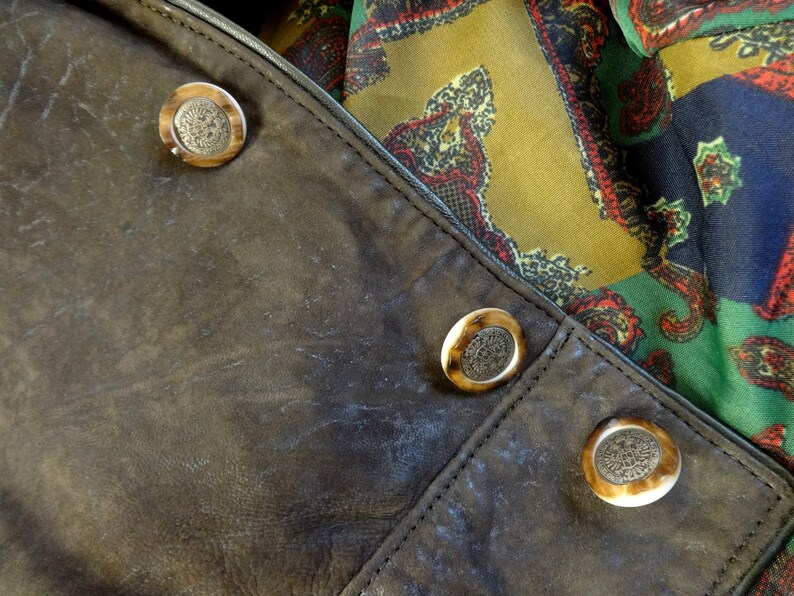 Puede incluir: Primer plano de una chaqueta de cuero marr&oacute;n con tres botones decorativos. Los botones son redondos y tienen un color marr&oacute;n claro con un centro marr&oacute;n oscuro.