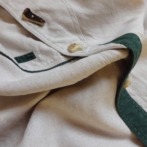 Pu&ograve; includere: Primo piano di un capo in lino beige con finiture verde scuro e bottoni decorativi. I bottoni sono realizzati in un materiale chiaro con un aspetto strutturato unico. Il capo ha un design classico e su misura.