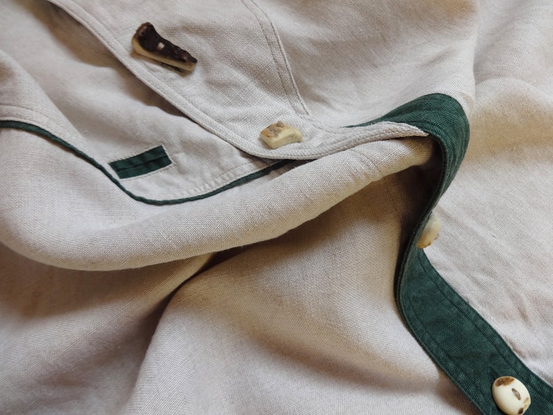 Pu&ograve; includere: Primo piano di un capo in lino beige con finiture verde scuro e bottoni decorativi. I bottoni sono realizzati in un materiale chiaro con un aspetto strutturato unico. Il capo ha un design classico e su misura.