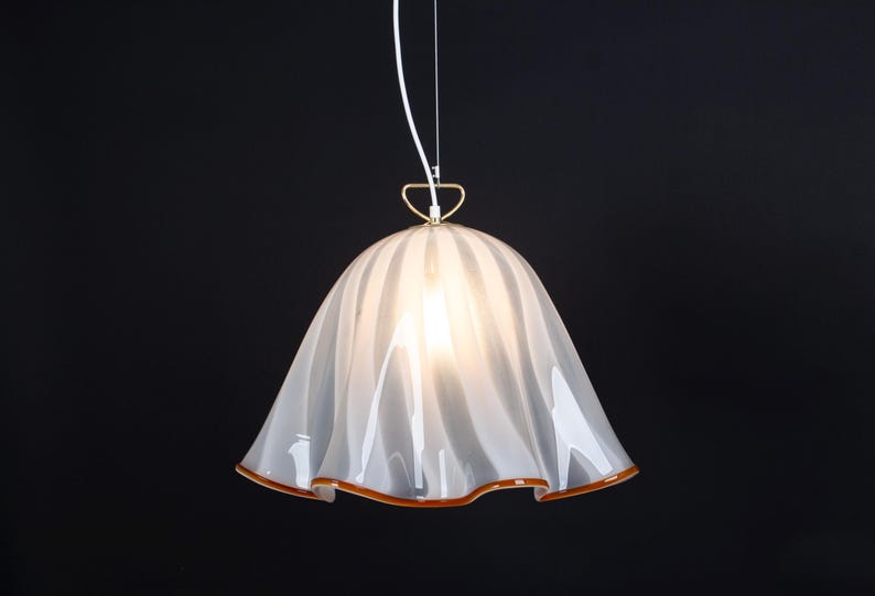 May include: A white glass pendant light with a ruffled, wavy design. The light has a gold metal ring at the top and a white cord.