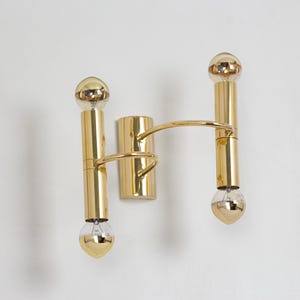 May include: A gold-toned wall sconce with two vertical light fixtures and a connecting curved arm. Each fixture has a bulb at the top and bottom. The sconce is mounted on a wall, showcasing a modern lighting design.