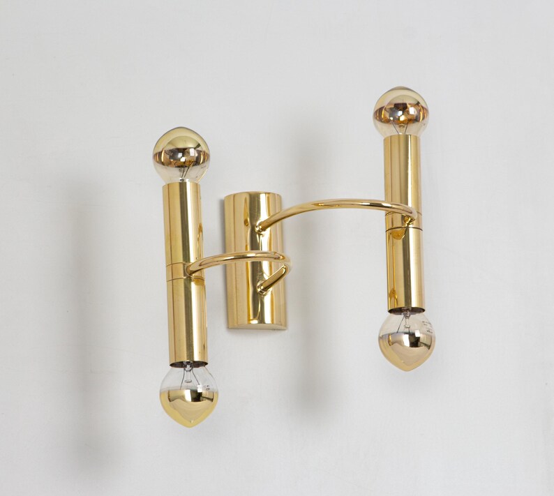 May include: A gold-toned wall sconce with two vertical light fixtures and a connecting curved arm. Each fixture has a bulb at the top and bottom. The sconce is mounted on a wall, showcasing a modern lighting design.