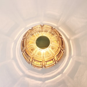 Könnte beinhalten: Eine runde, bernsteinfarbene Glas-Deckenleuchte mit geometrischem Muster. Die Leuchte hat eine goldfarbene Mitte.