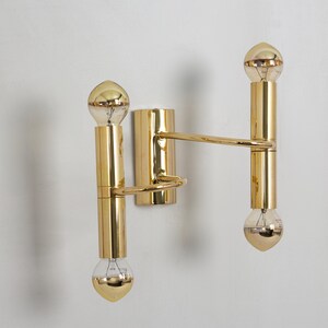 May include: A modern, gold-toned wall sconce with two light fixtures. The sconce features a rectangular base, cylindrical arms, and globe-shaped bulbs. The design is minimalist and elegant, suitable for contemporary interiors.