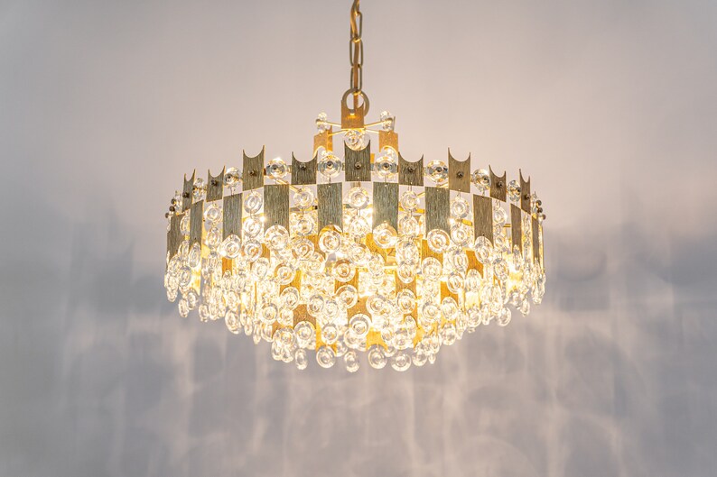 Peut inclure: Un lustre dor&eacute; avec un cadre circulaire et des gouttes de cristal en cascade. Le lustre a un design g&eacute;om&eacute;trique avec une s&eacute;rie de barres verticales.