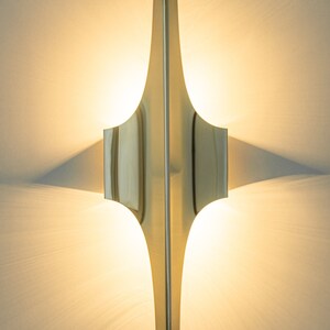 May include: A gold-toned wall sconce with a geometric design. The sconce is illuminated and casts a warm glow on the wall.