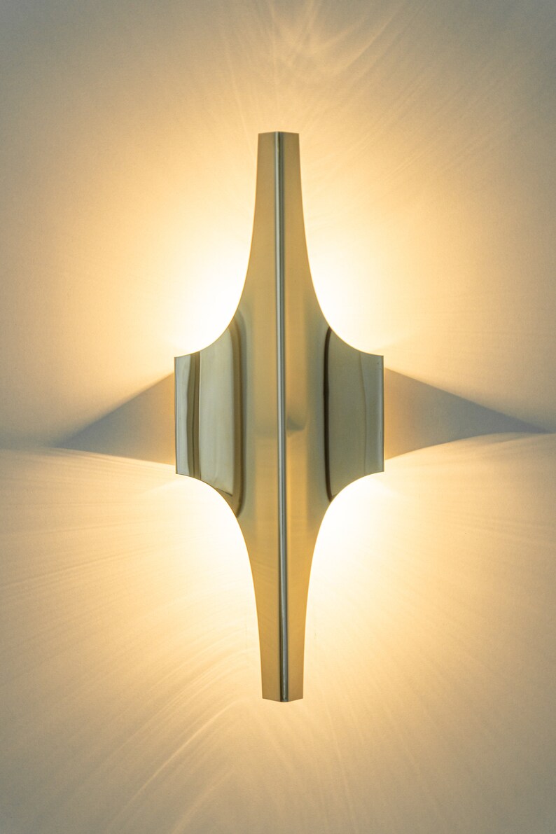 May include: A gold-toned wall sconce with a geometric design. The sconce is illuminated and casts a warm glow on the wall.
