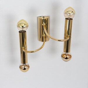 May include: A gold-colored wall sconce with two light bulbs. The sconce has a central mounting plate and two curved arms that hold the light bulbs. The light bulbs are round and have a gold-colored finish. The sconce is mounted on a white wall.
