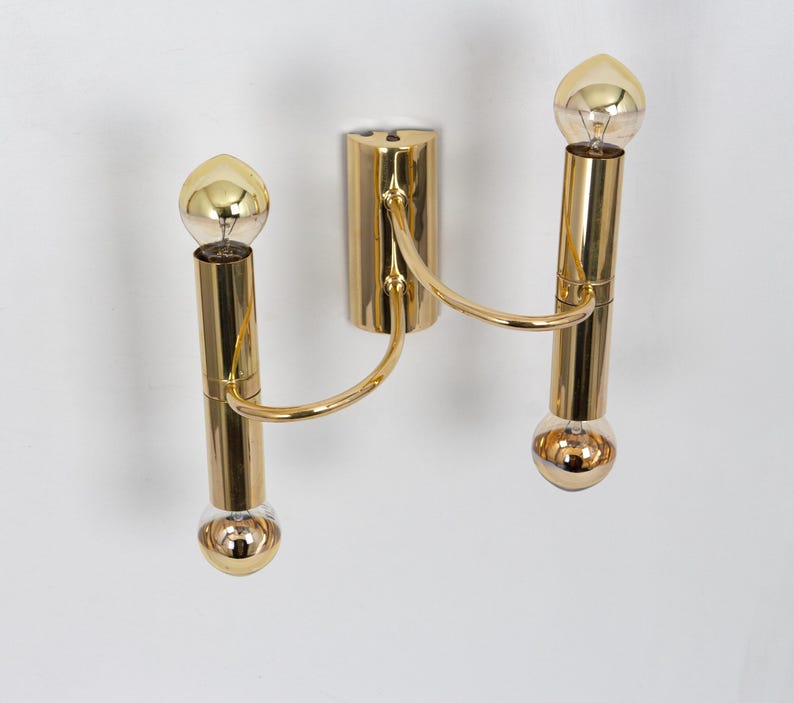 May include: A gold-colored wall sconce with two light bulbs. The sconce has a central mounting plate and two curved arms that hold the light bulbs. The light bulbs are round and have a gold-colored finish. The sconce is mounted on a white wall.