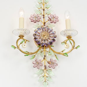 Puede incluir: Una l&aacute;mpara de pared dorada con un dise&ntilde;o floral. La l&aacute;mpara tiene dos bombillas de estilo vela y presenta flores y hojas de vidrio rosa, morado y verde.