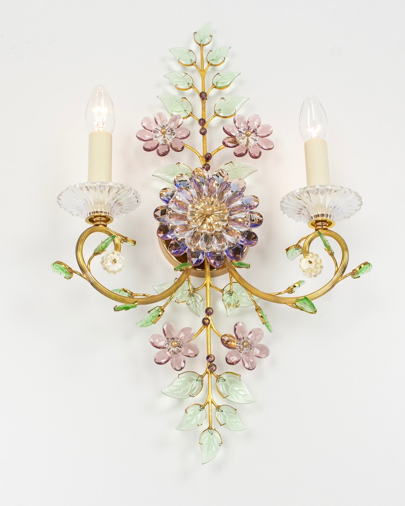 Puede incluir: Una l&aacute;mpara de pared dorada con un dise&ntilde;o floral. La l&aacute;mpara tiene dos bombillas de estilo vela y presenta flores y hojas de vidrio rosa, morado y verde.