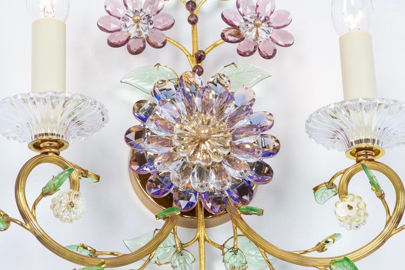 Puede incluir: Una l&aacute;mpara de pared dorada con dos bombillas de estilo vela y flores de cristal. Las flores son en tonos de rosa, morado y verde.