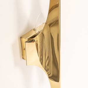 May include: A gold-toned wall sconce with a curved, geometric design. The sconce has a single light bulb and a clear glass shade.