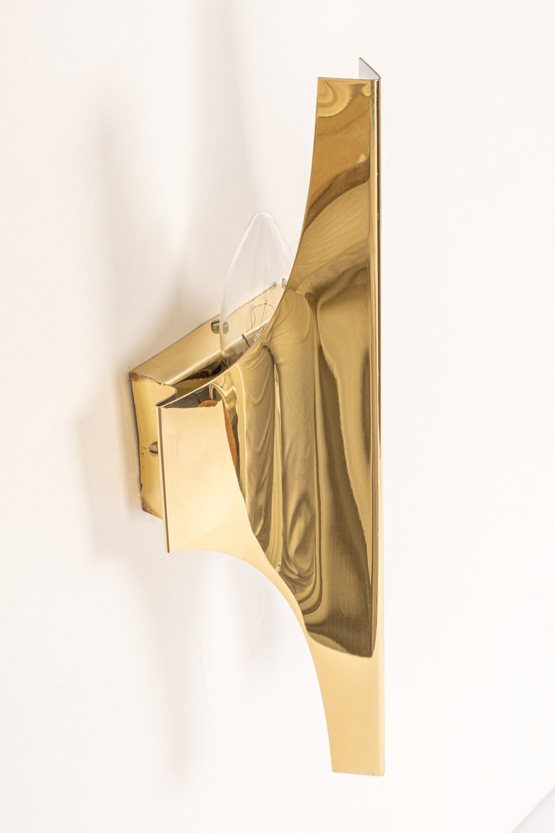 May include: A gold-toned wall sconce with a curved, geometric design. The sconce has a single light bulb and a clear glass shade.