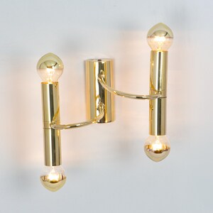 May include: A gold-colored wall sconce with two vertical light fixtures. Each fixture has a bulb at the top and bottom, connected by a curved arm to a central mounting plate. The bulbs are illuminated, casting a warm glow.