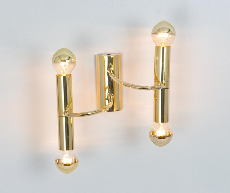 May include: A gold-colored wall sconce with two vertical light fixtures. Each fixture has a bulb at the top and bottom, connected by a curved arm to a central mounting plate. The bulbs are illuminated, casting a warm glow.