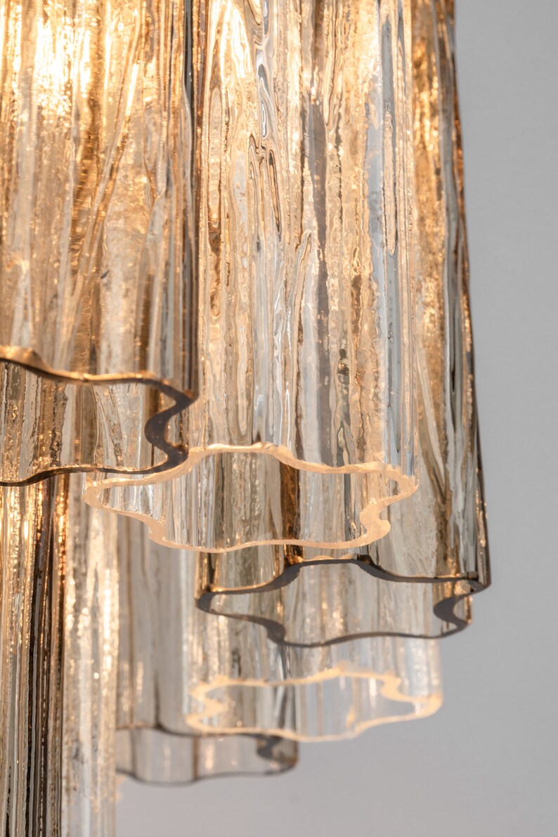 May include: A close-up of a multi-tiered glass chandelier with a rippled, textured surface. The glass is clear with a slight amber hue. The chandelier has a modern design and would be a stylish addition to any home.
