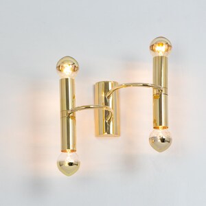 May include: A gold-colored wall sconce with two vertical light fixtures, each holding a bulb at the top and bottom. A curved arm connects the two fixtures. The sconce is mounted on a white wall.
