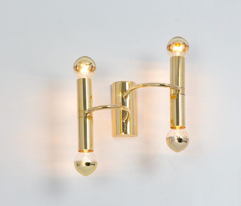 May include: A gold-colored wall sconce with two vertical light fixtures, each holding a bulb at the top and bottom. A curved arm connects the two fixtures. The sconce is mounted on a white wall.