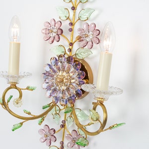 Puede incluir: Una l&aacute;mpara de pared dorada con dos bombillas de estilo vela. La l&aacute;mpara est&aacute; decorada con flores de vidrio morado, rosa y verde.
