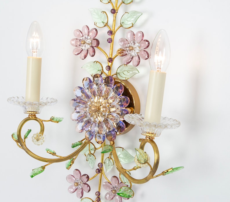 Puede incluir: Una l&aacute;mpara de pared dorada con dos bombillas de estilo vela. La l&aacute;mpara est&aacute; decorada con flores de vidrio morado, rosa y verde.