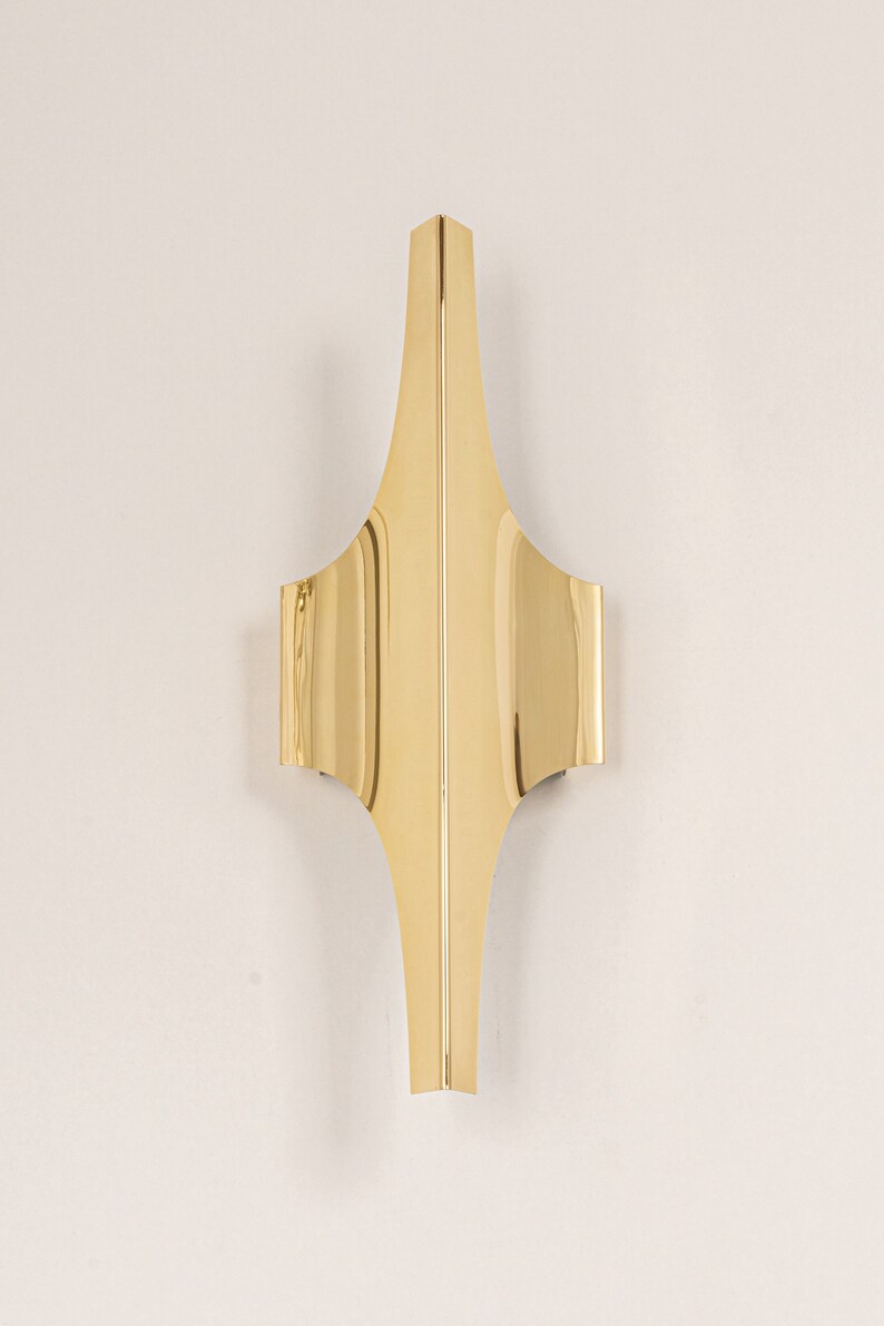 May include: A gold-coloured metal wall sconce with a geometric design. The sconce has a pointed top and a curved bottom.