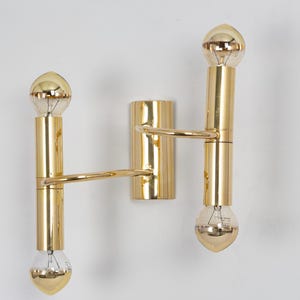 May include: A gold-colored, double-bulb wall sconce. The sconce features two vertical cylindrical sections connected by a horizontal arm. Each section holds a teardrop-shaped light bulb. The fixture is mounted on a rectangular base.
