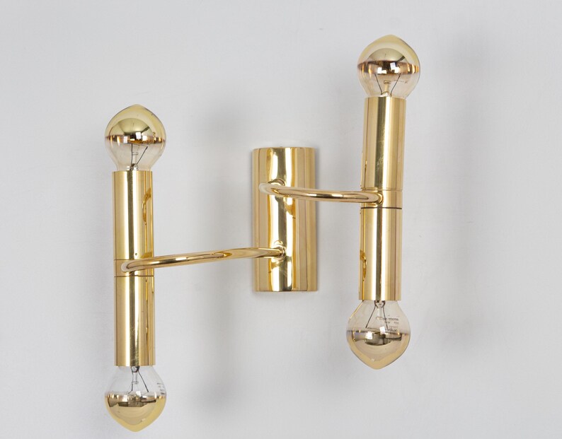 May include: A gold-colored, double-bulb wall sconce. The sconce features two vertical cylindrical sections connected by a horizontal arm. Each section holds a teardrop-shaped light bulb. The fixture is mounted on a rectangular base.