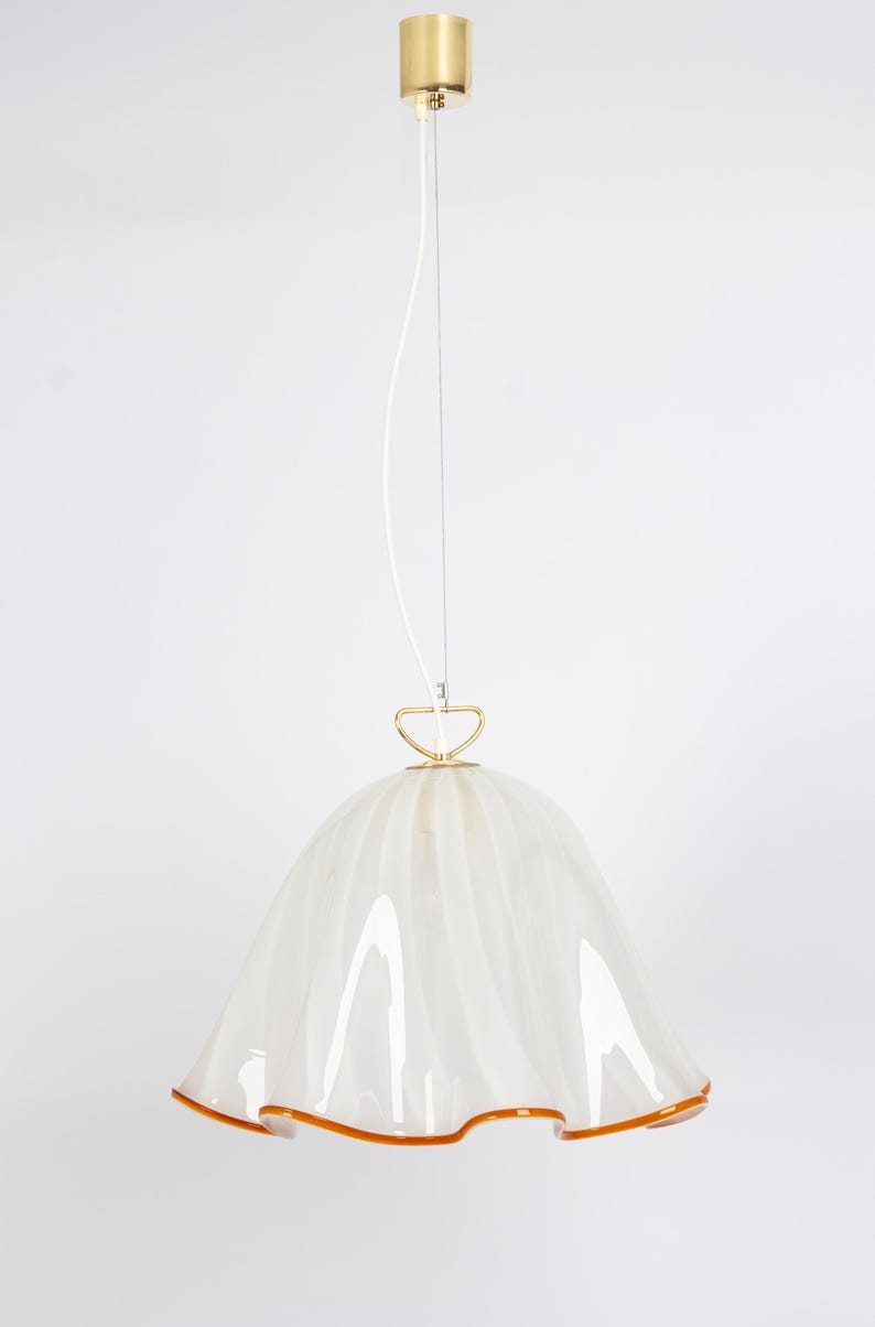 May include: White glass pendant light with a ruffled edge and a gold metal top. The light has a white cord and a gold metal ring.