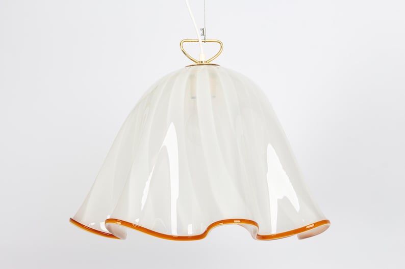 May include: A white glass pendant light with a gold metal canopy and a ruffled, scalloped edge. The light has a warm, amber-colored trim.