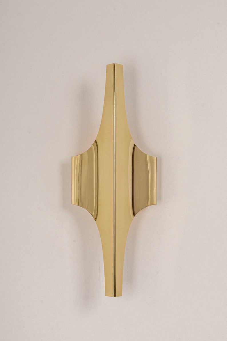 May include: A gold metal wall sconce with a geometric design. The sconce has a pointed top and a wide base.