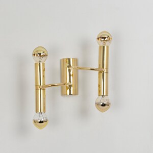 May include: A gold-colored wall sconce with two vertical light fixtures and a central mounting plate. Each light fixture has a cylindrical body with a bulb at each end. The sconce has a minimalist design and a polished finish.