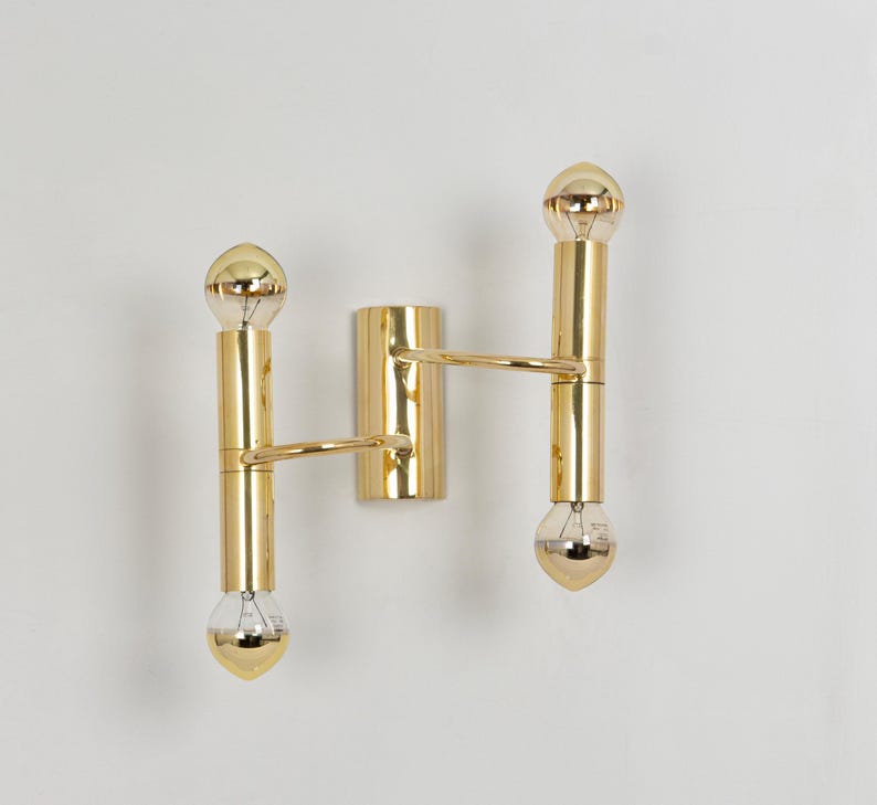 May include: A gold-colored wall sconce with two vertical light fixtures and a central mounting plate. Each light fixture has a cylindrical body with a bulb at each end. The sconce has a minimalist design and a polished finish.