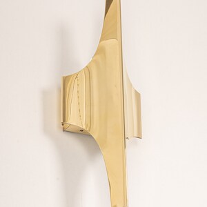 May include: A gold-toned wall sconce with a geometric design. The sconce has a curved shape and a sharp point at the top.