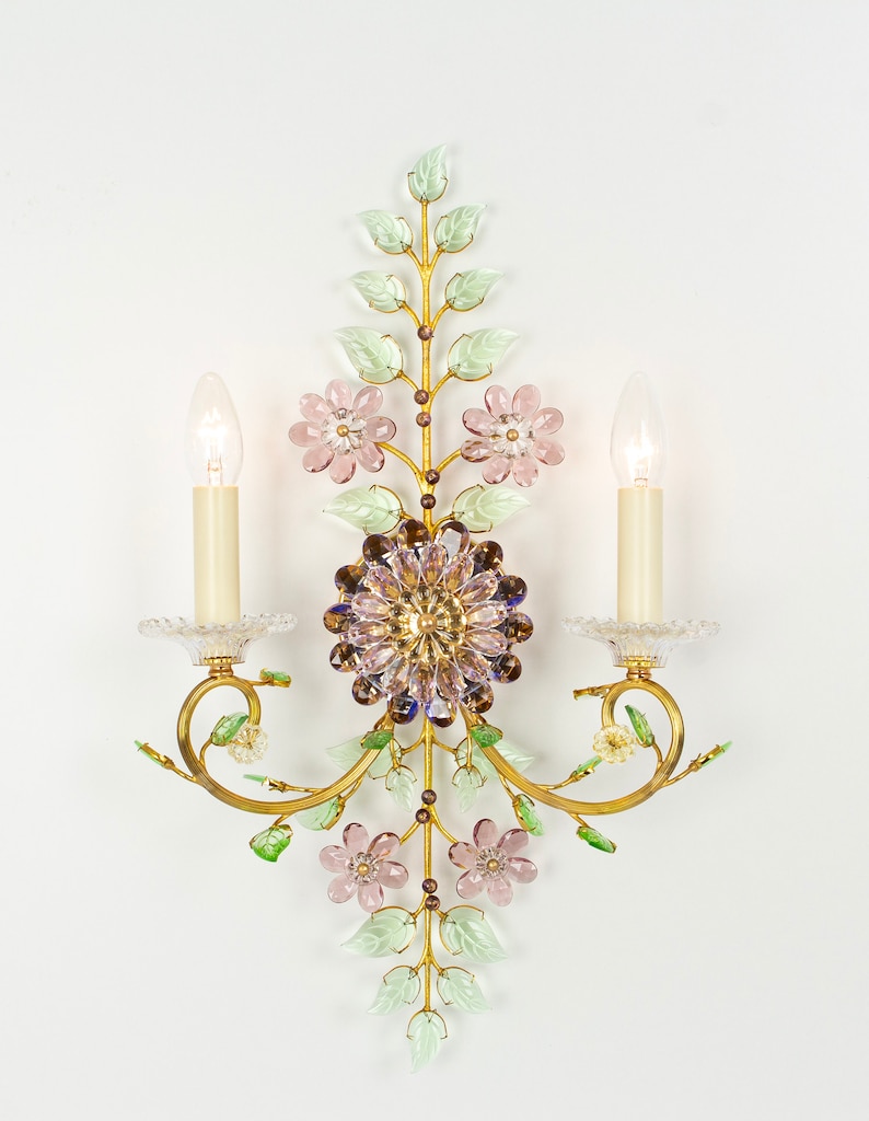 Puede incluir: Una l&aacute;mpara de pared dorada con un dise&ntilde;o floral. La l&aacute;mpara tiene dos bombillas de estilo vela y presenta flores y hojas de vidrio rosa y verde.