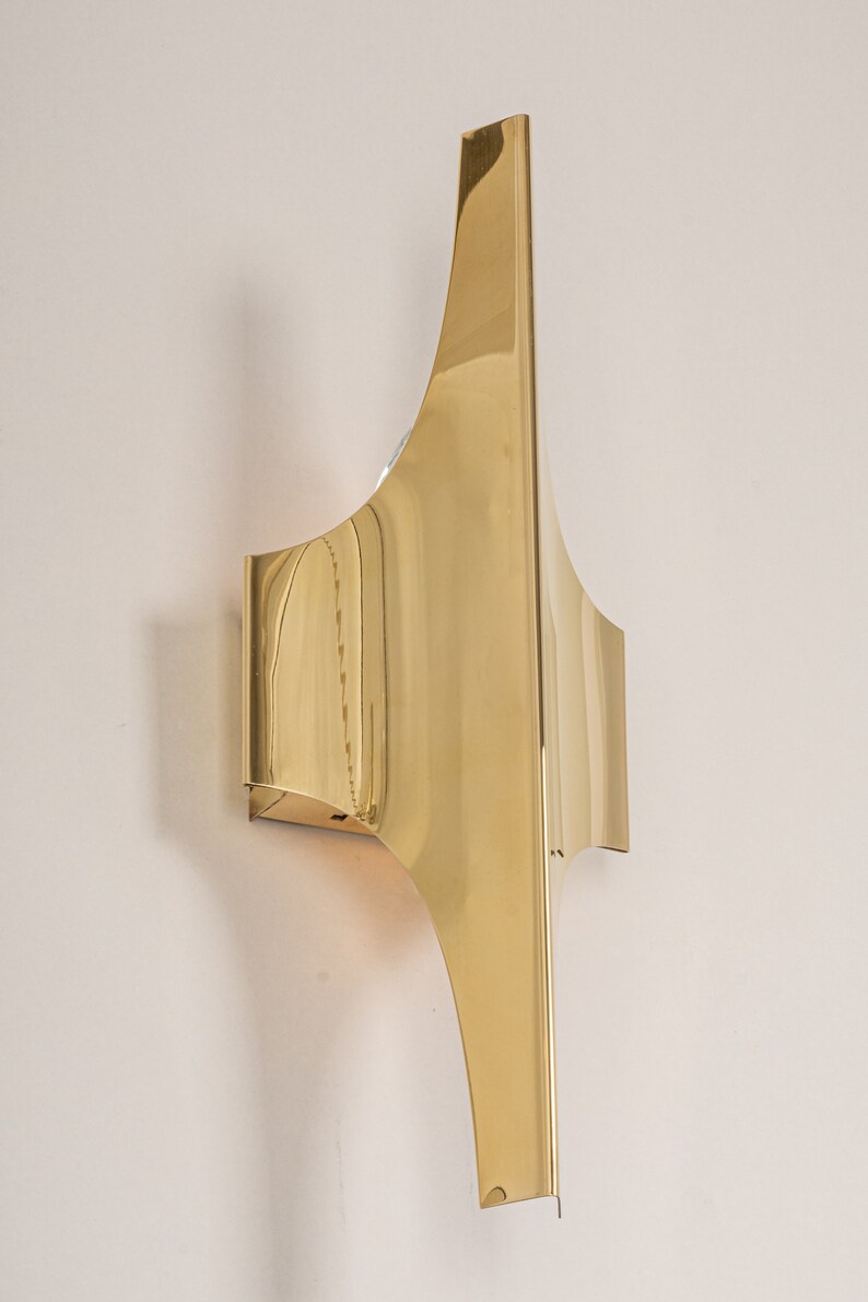 May include: A gold-toned metal wall sconce with a unique, geometric design. The sconce has a curved, angular shape and a polished finish.