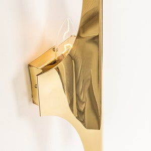 May include: A gold-toned wall sconce with a curved, geometric design. The sconce has a single light bulb.