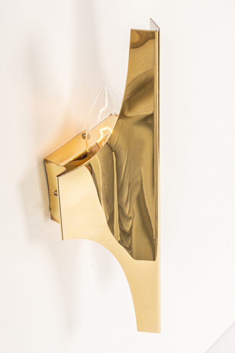 May include: A gold-toned wall sconce with a curved, geometric design. The sconce has a single light bulb.