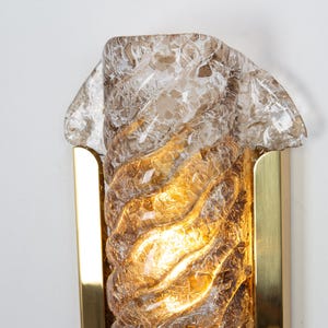 May include: A gold wall sconce with a clear, textured glass shade. The glass has a swirling pattern and is illuminated from within.