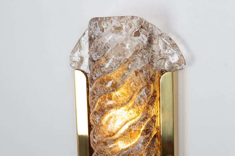 May include: A gold wall sconce with a clear, textured glass shade. The glass has a swirling pattern and is illuminated from within.