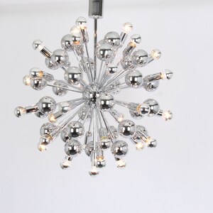 Op de afbeelding: Een chromen Sputnik-kroonluchter met meerdere lampen. De kroonluchter heeft een ronde vorm met veel armen die naar buiten uitstrekken.